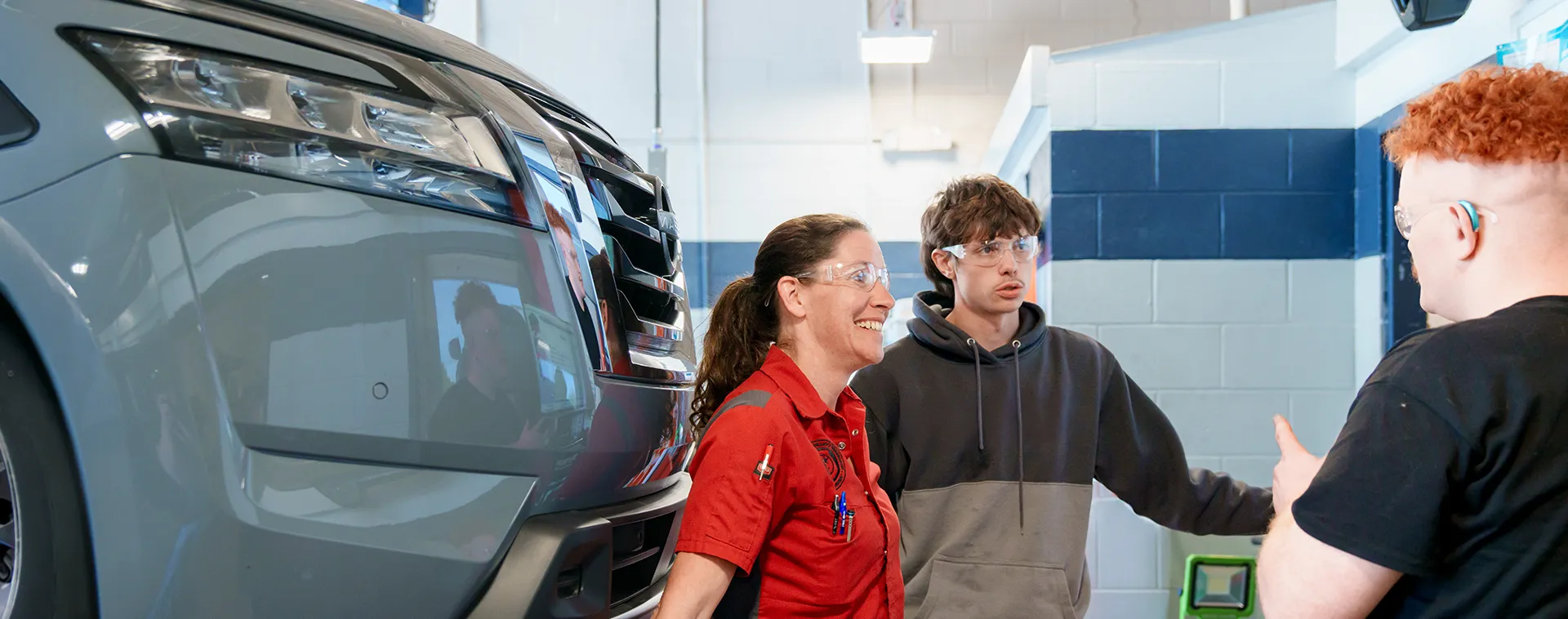 Instructor and two students wearing safety glasses stand beside a vehicle in an automotive training lab.