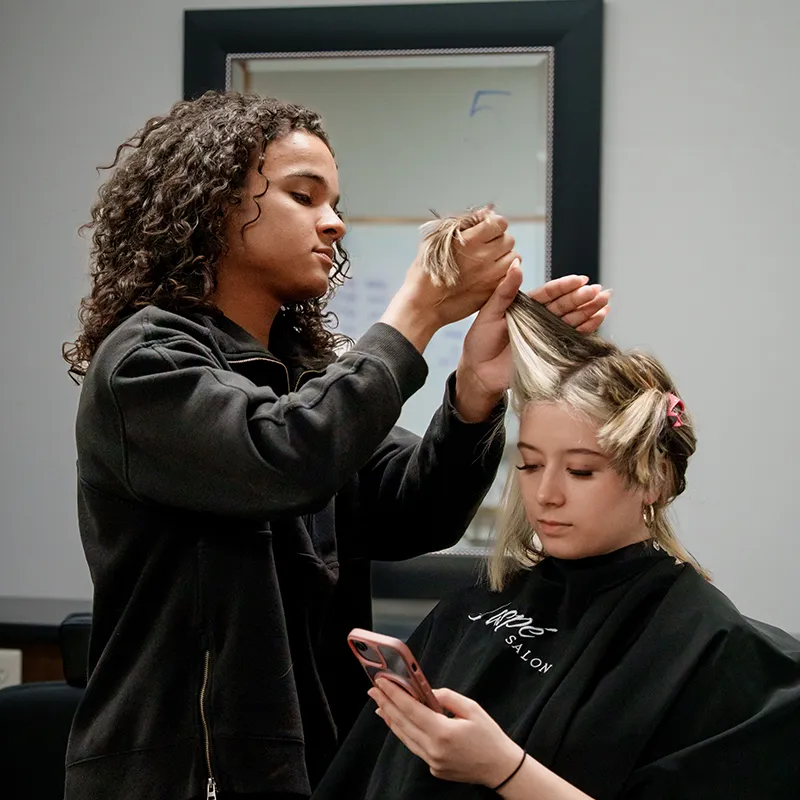 Student sections and styles hair for a seated client in a cosmetology training salon.