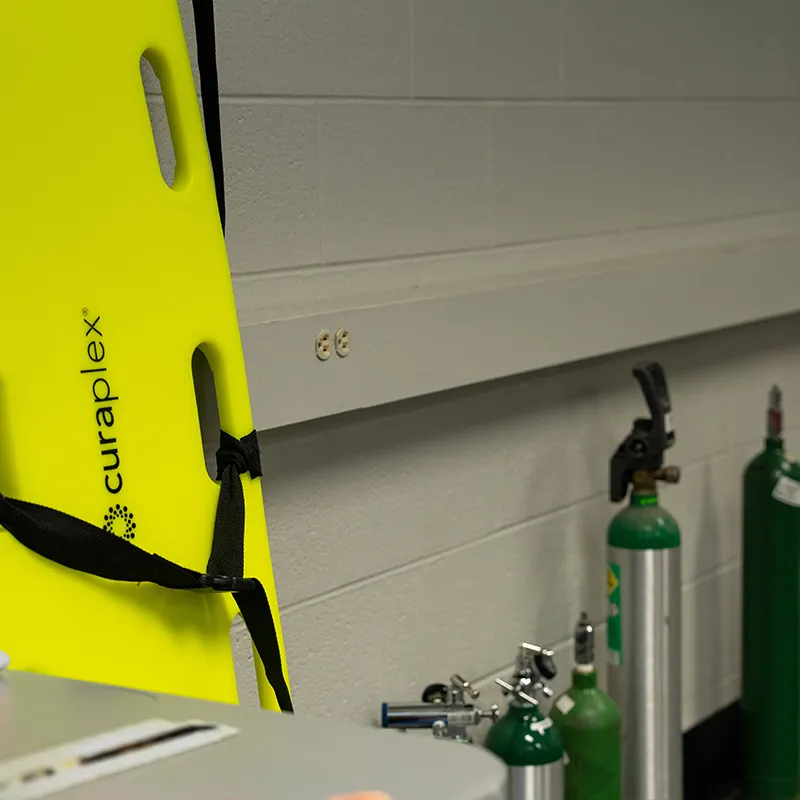 Bright yellow spine board mounted beside oxygen cylinders and medical training equipment.