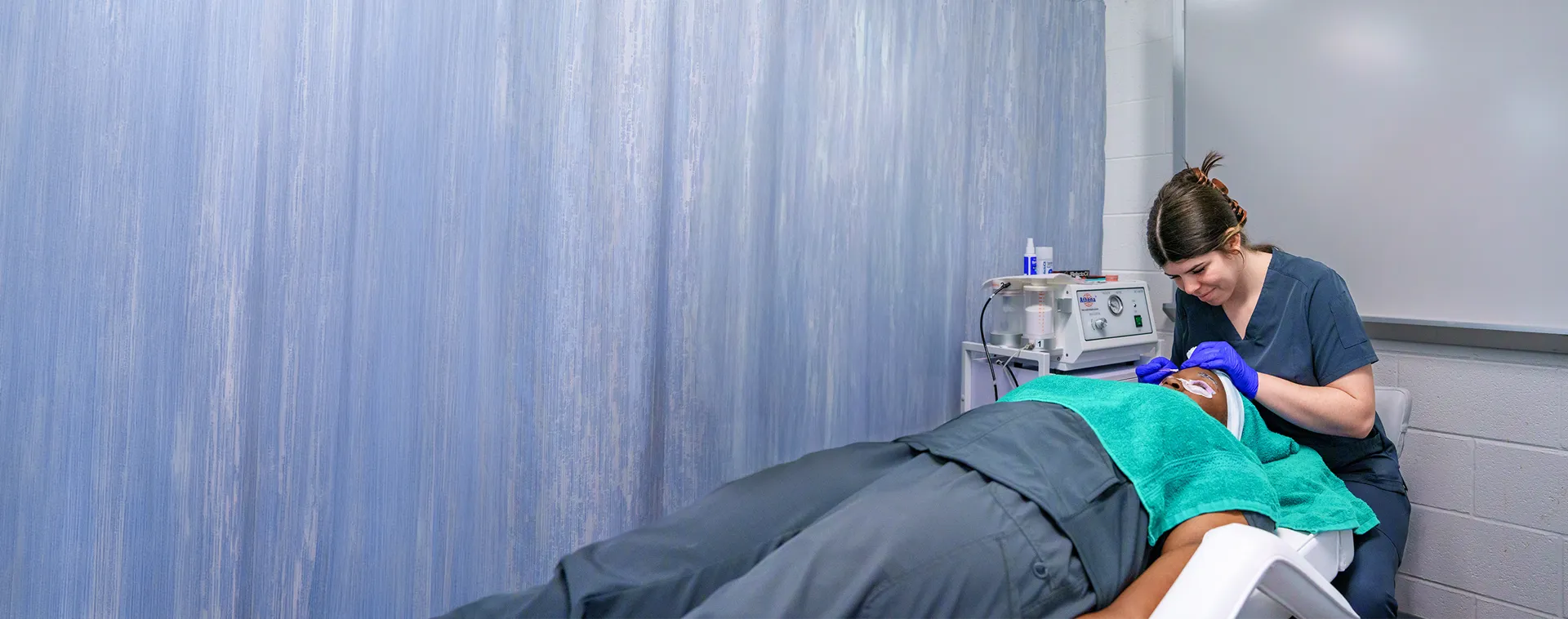 Person in scrubs performs a facial treatment on a client reclining on a treatment bed in a skincare lab.