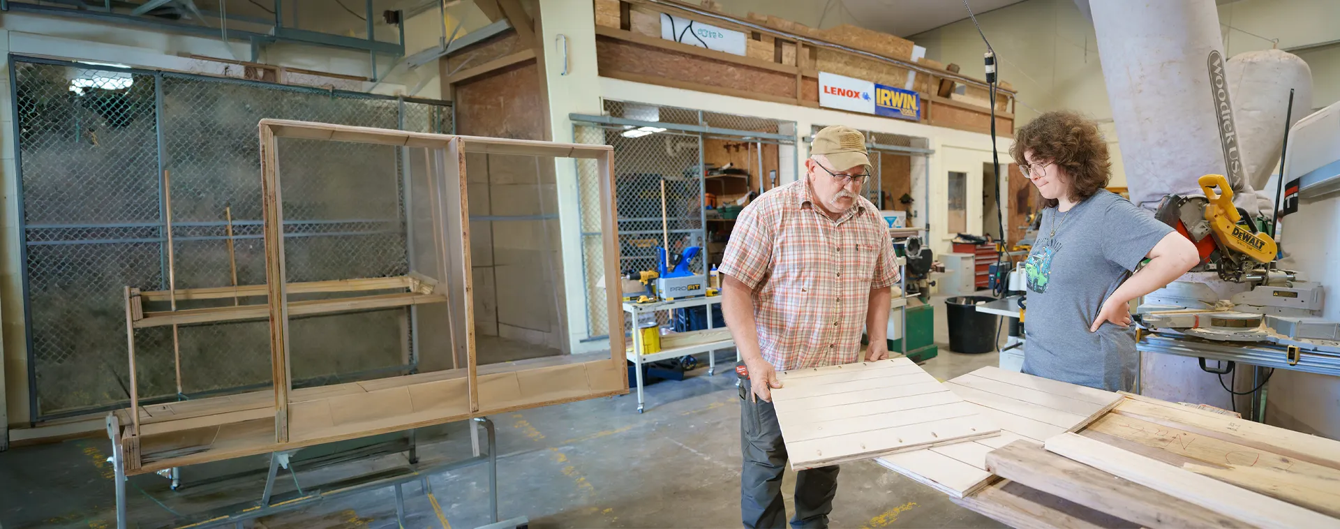 Instructor and student review wood panels beside a framed structure in a construction training workshop.