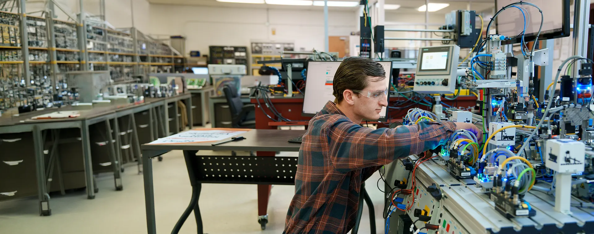 Person wearing safety glasses adjusts wiring on an automated electronics training system in a lab filled with equipment.
