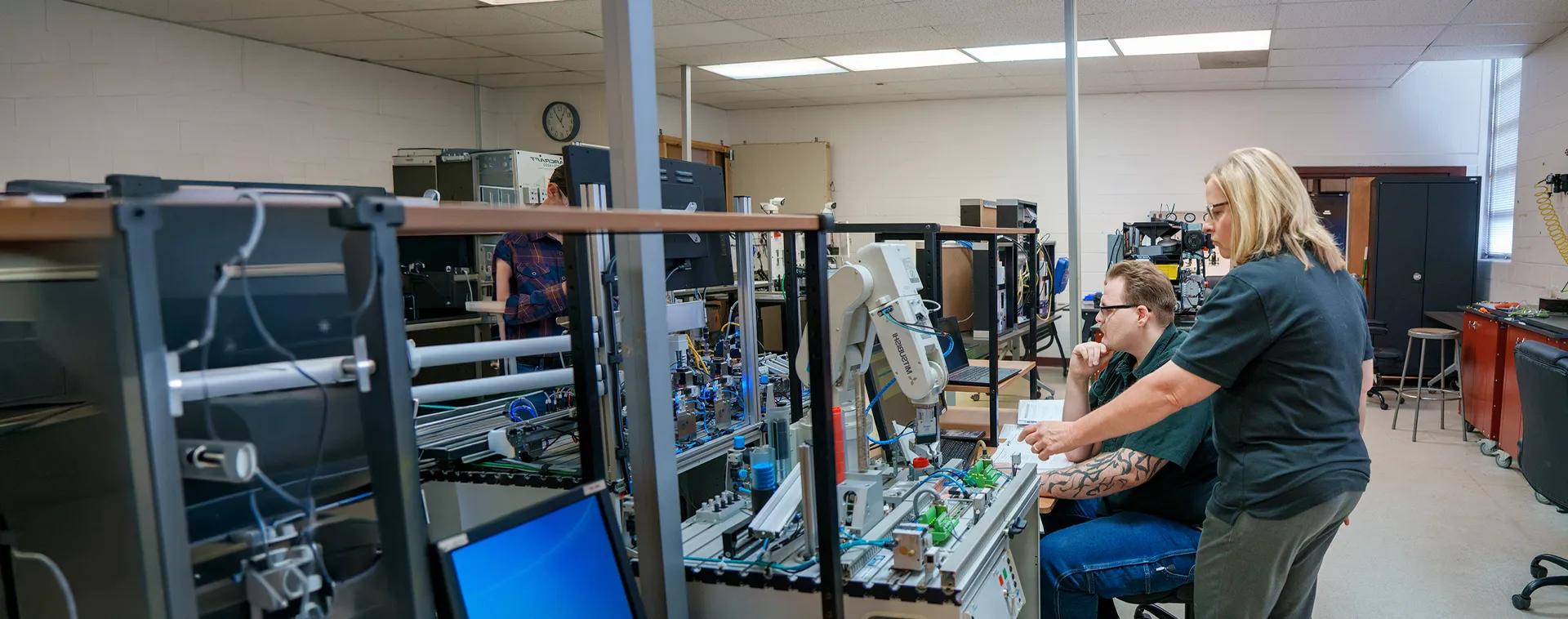 Instructor and student review an automated manufacturing training system with robotic equipment in an electronics lab.