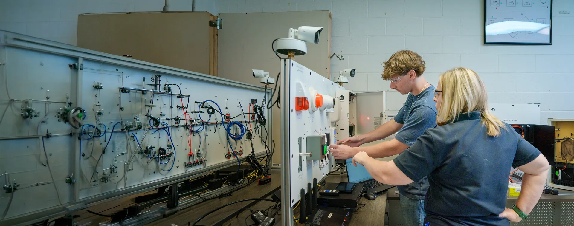 Instructor and student operating an electronics training panel with wiring, controls, and monitoring equipment in a lab.