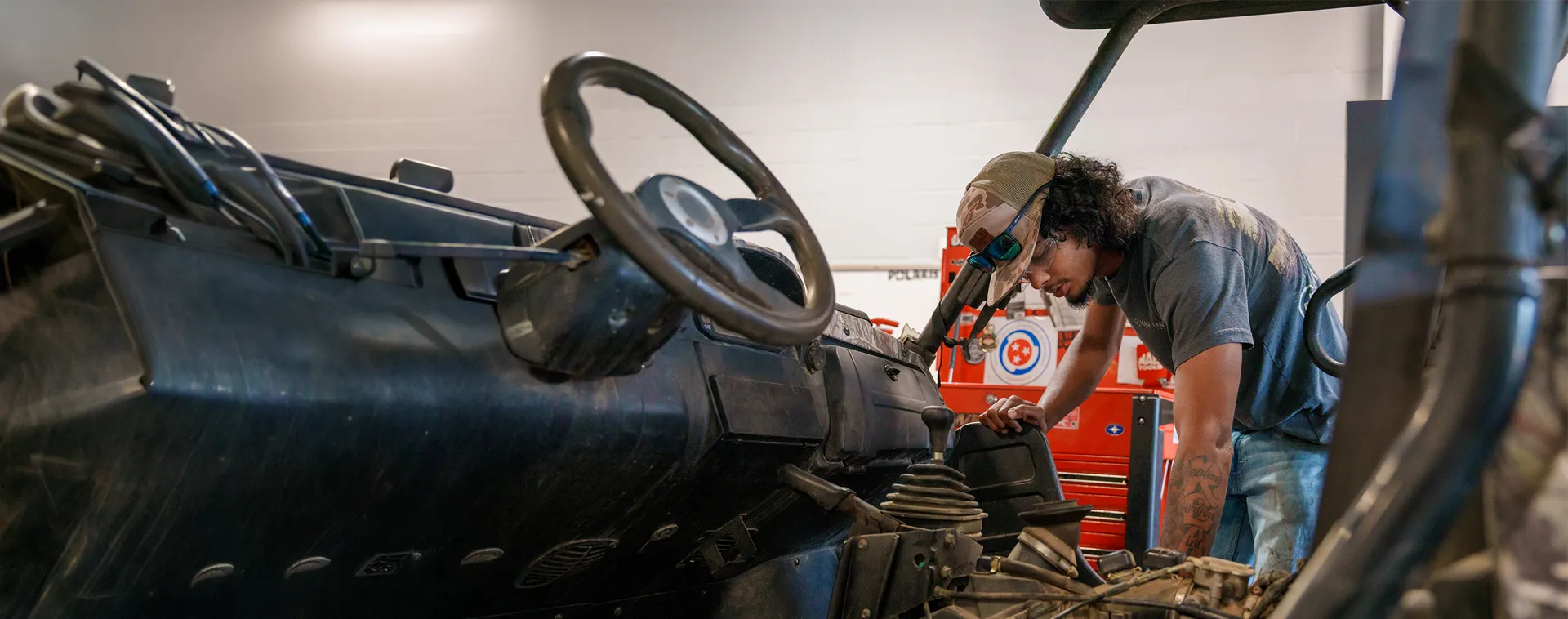 Person leaning into an off-road utility vehicle, inspecting components inside a workshop with tool cabinets in back.