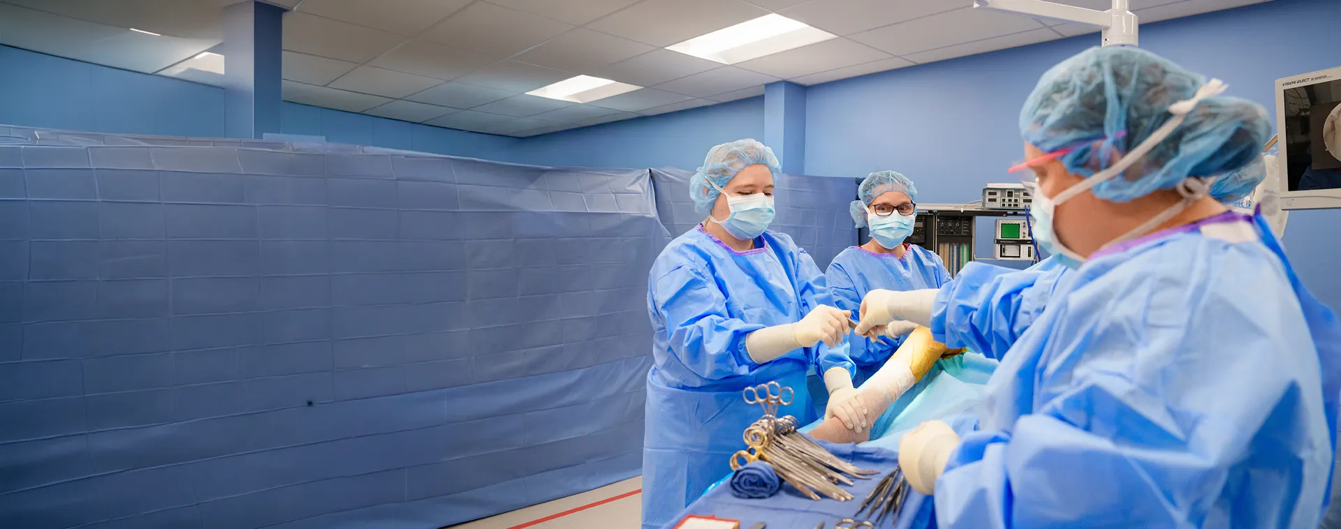 Surgical technology students in blue sterile gowns and masks passing instruments during a simulated procedure.