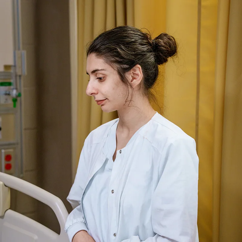 Student in a clinical uniform stands beside a hospital bed in a patient care training room.