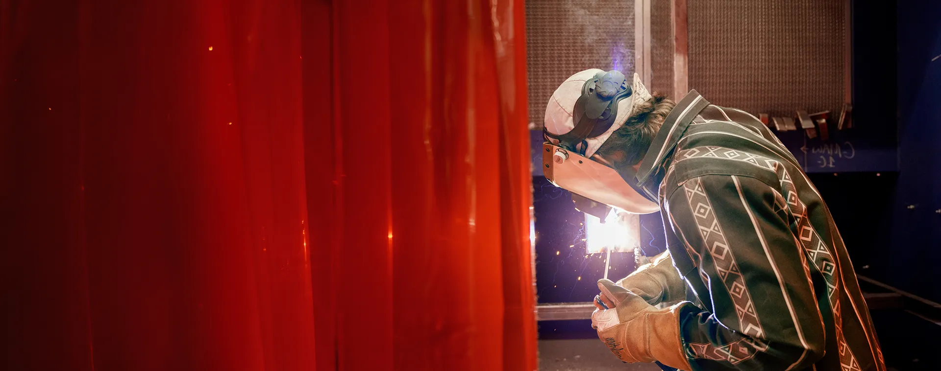 Student wearing a welding helmet and gloves performs a weld behind a red welding curtain as sparks glow.
