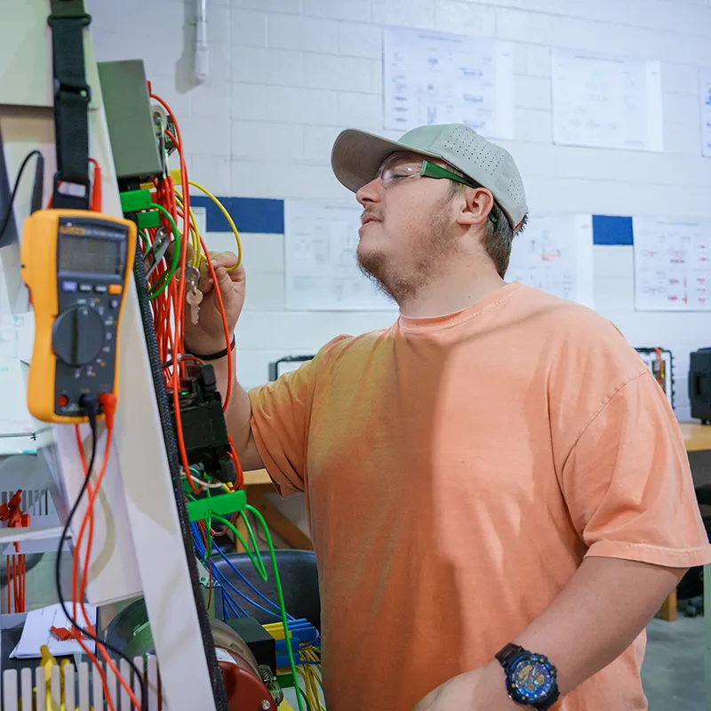 Student inspects wiring and electrical components on a training panel with multimeter leads and cables.