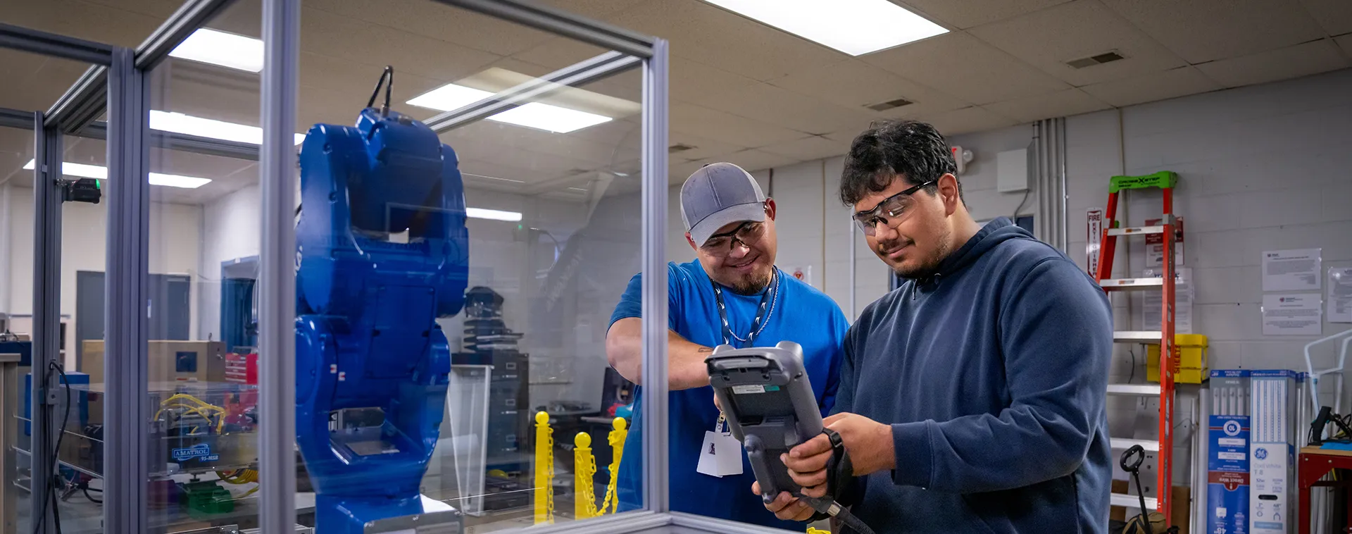 Instructor and student review a handheld control device beside an industrial robot enclosed in a training cell.