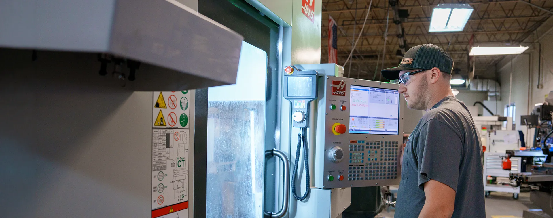 Student wearing safety glasses operates a CNC machine control panel in a manufacturing lab.