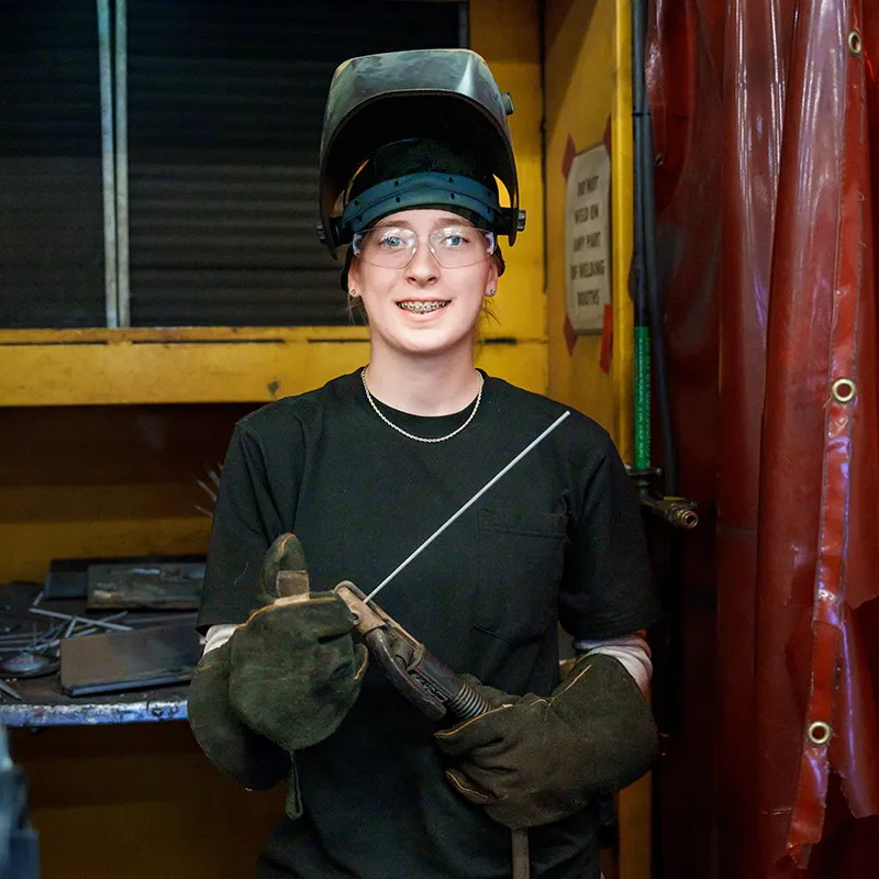 Welding student wearing a raised helmet, safety glasses, and gloves holds a welding electrode in a shop.