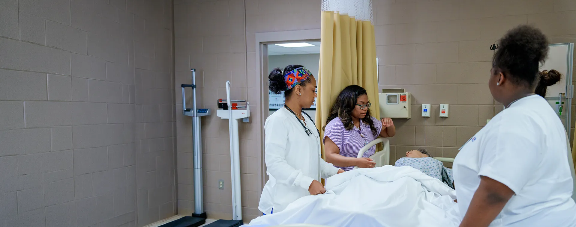 Students and an instructor provide care to a patient mannequin in a nursing skills lab with hospital equipment.