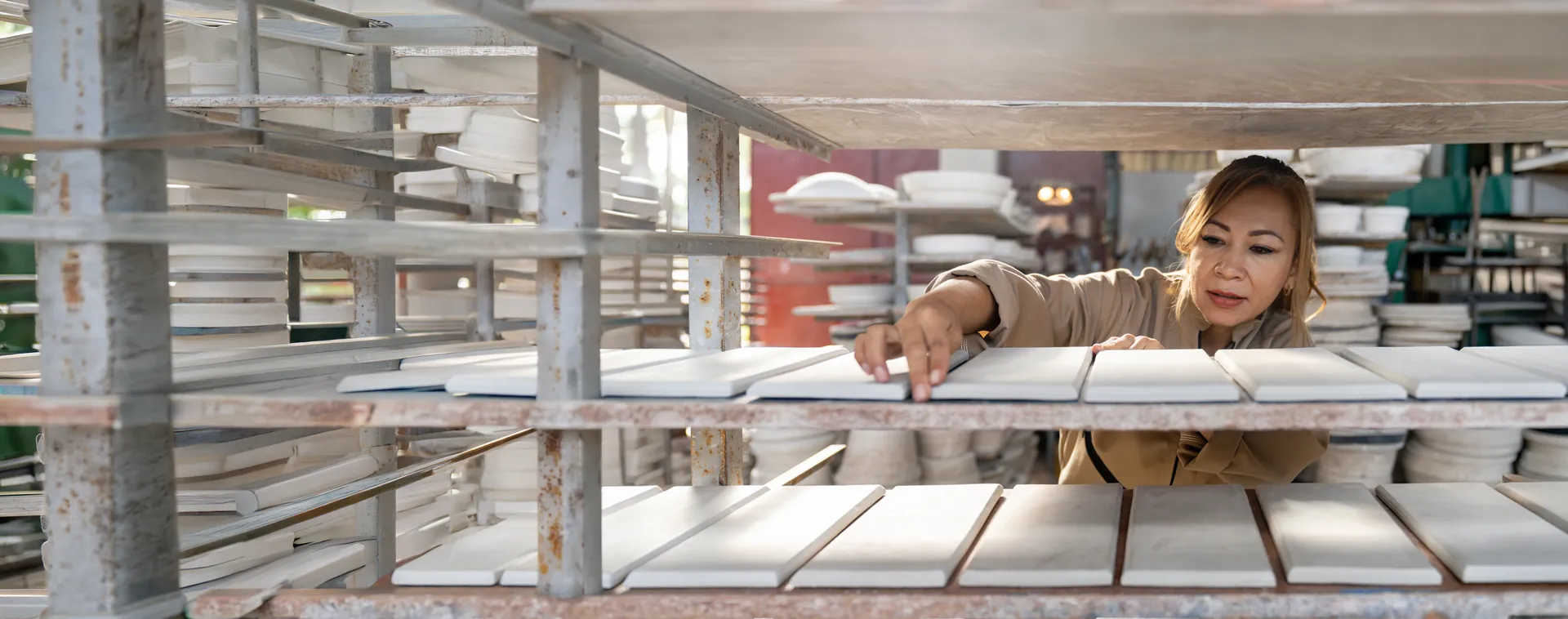 Person arranging white ceramic tiles on metal drying racks in a workshop with shelves of tile pieces in the background.