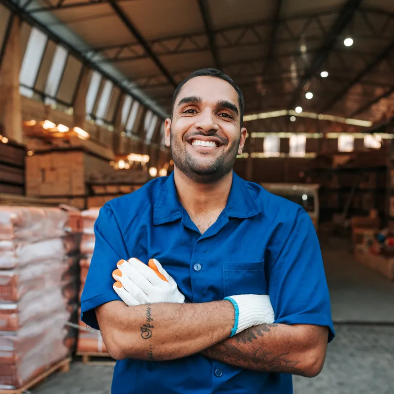 Worker in blue work shirt and gloves standing in a warehouse with stacked materials and industrial shelving behind.