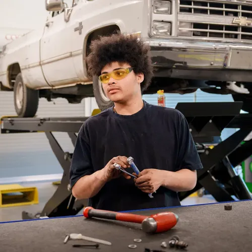 Student wearing safety glasses holds hand tools at a workbench with a pickup truck raised on a vehicle lift behind.