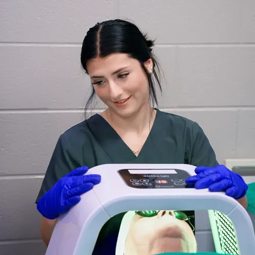 Person in scrubs and gloves operates a facial treatment device above a client during a skincare procedure.