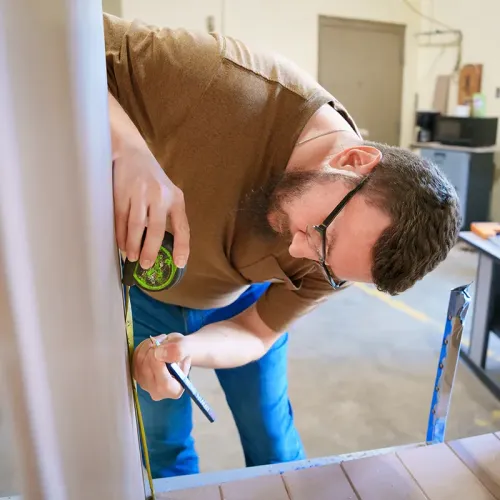 Student measures and marks a panel with a tape measure and pencil in a construction workshop.