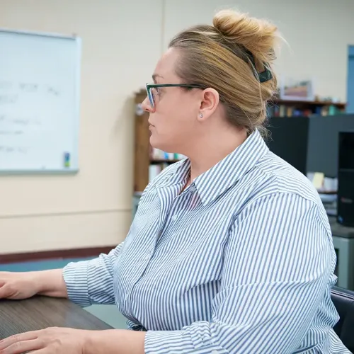 Person seated at a computer workstation in a CAD classroom with whiteboard and bookshelves in the background.