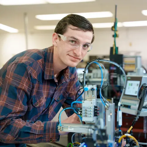 Person wearing safety glasses leans over an electronics training system with cables, sensors, and control equipment.