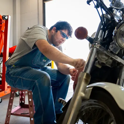 Person seated beside a motorcycle, using a hand tool near the front wheel in a bright workshop.
