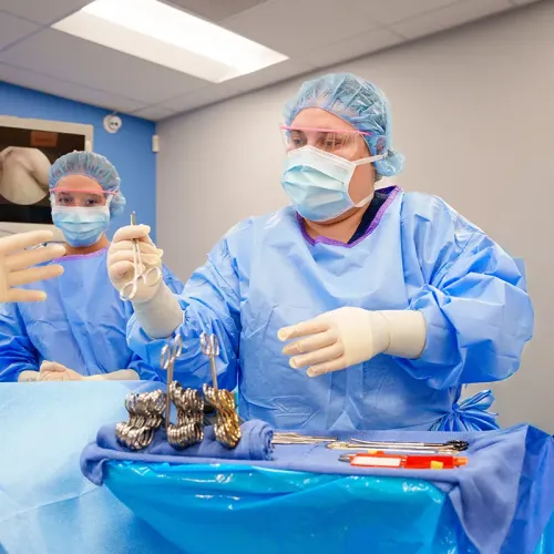 Surgical technology student in protective gown, mask, and face shield handing an instrument across a sterile field.
