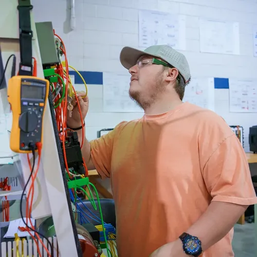 Student inspects wiring and electrical components on a training panel with multimeter leads and cables.