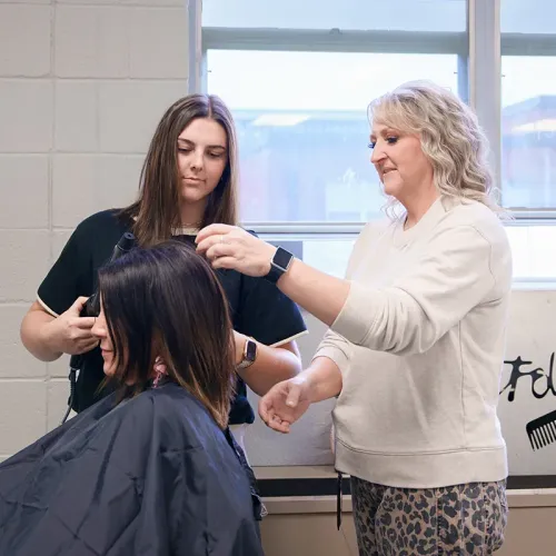 Instructor demonstrates hairstyling techniques while a student works with a client in a cosmetology classroom.