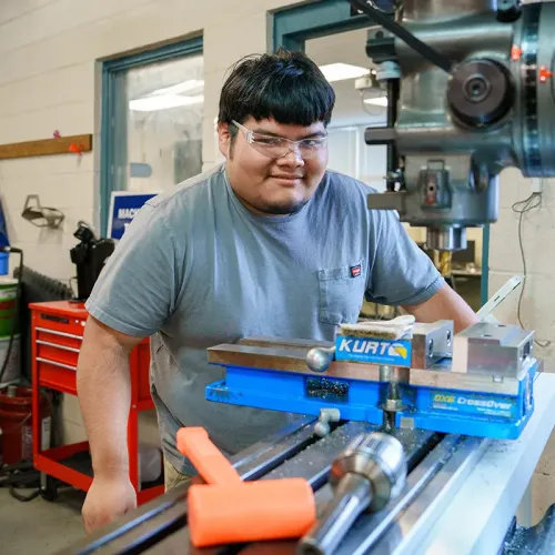 Student wearing safety glasses stands beside a milling machine with a vise and metal workpiece.