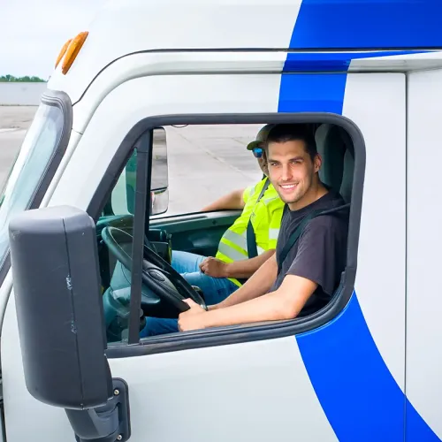 Student seated behind the wheel of a training truck with an instructor visible in the passenger seat.