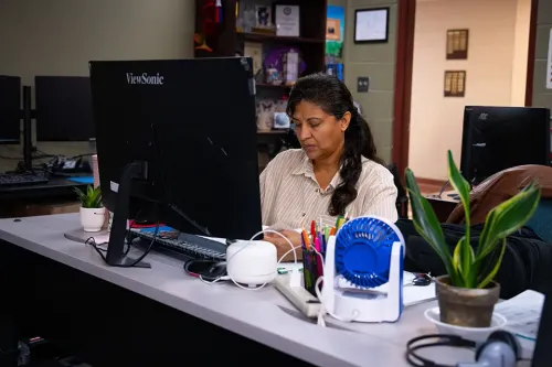 Person working at a desk with a computer monitor, desk fan, office supplies, plants, and shelving in the background.