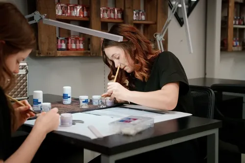 Student applies nail product with a brush at a manicure workstation with supplies and nail polish displays.