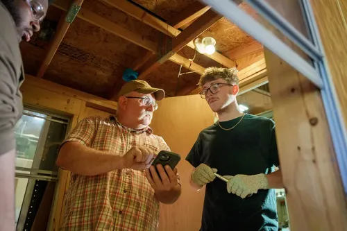 Instructor shows information on a phone to a student wearing work gloves inside a wood-framed training structure.