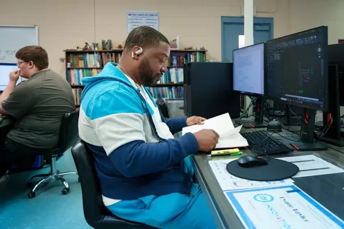 Student reviews printed materials beside dual monitors showing technical drawings and CAD software.