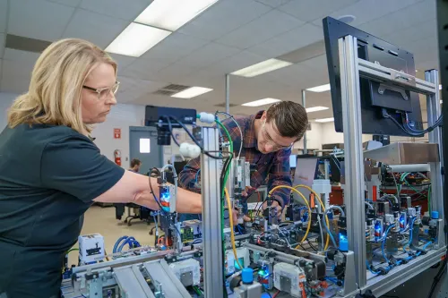 Two people wearing safety glasses work on an industrial automation training machine with monitors and wiring.