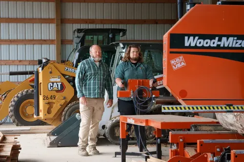 Two people stand beside a sawmill machine and skid steer loader inside a forestry training facility.