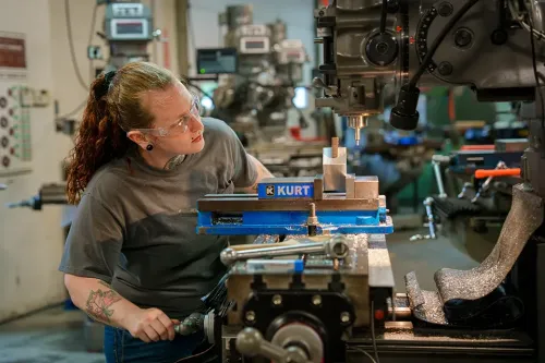 Person wearing safety glasses inspects a milling machine while adjusting controls in a machine shop.