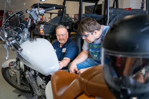 Two people examining a white motorcycle in a workshop, with utility vehicles and a helmet visible nearby.