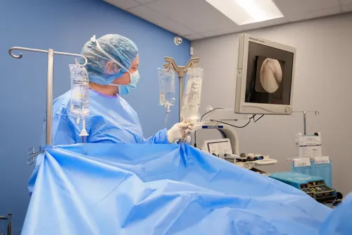 Student in surgical gown and mask monitors equipment and IV bags beside a draped operating table.