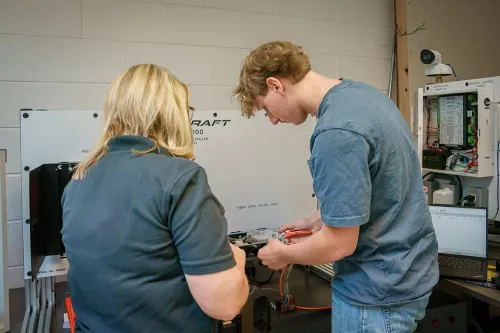 Instructor and student connecting wiring components at a fiber optic splice case workstation with a laptop nearby.