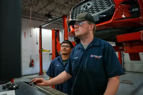 Student wearing safety glasses holds hand tools at a workbench with a pickup truck raised on a vehicle lift behind.